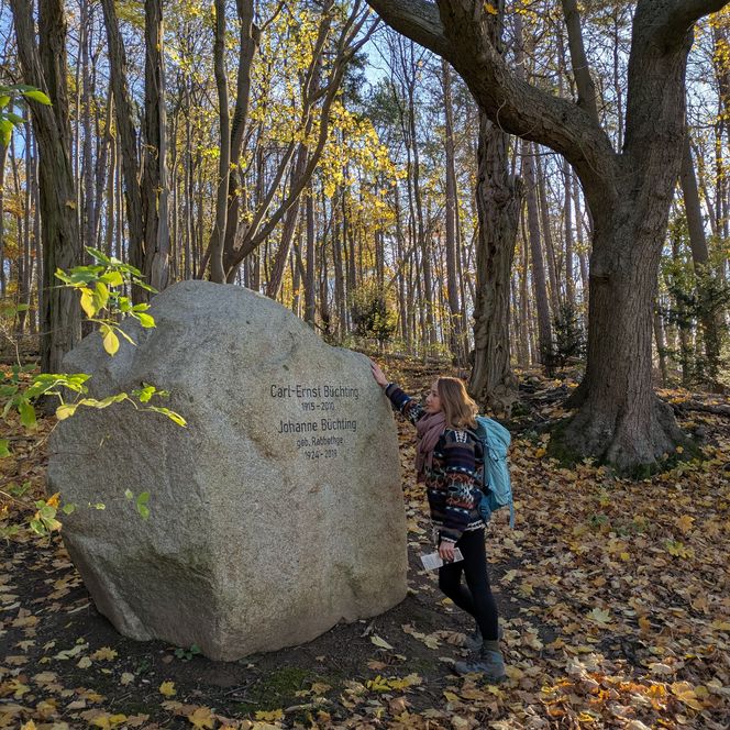 Frau mit Rucksack steht im herbstlichen Wald am Domeier Gedenkstein, umgeben von buntem Laub.
