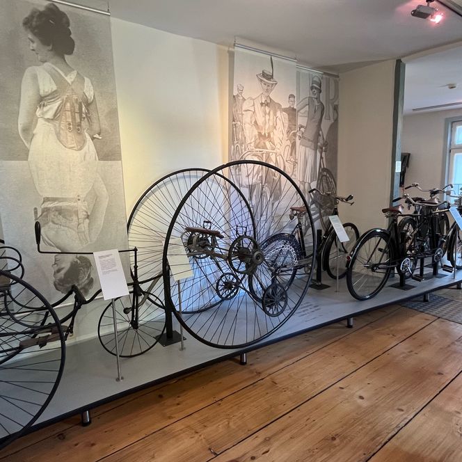 Historische Fahrräder, darunter ein Hochrad, ausgestellt im Museum vor großformatigen Wandbildern.