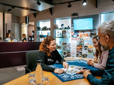 Mitarbeiterin berät zwei Gäste an einem Tisch in der Tourist-Information Einbeck.