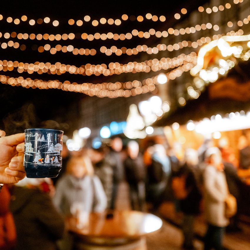Hand, die eine Tasse Glühwein hält im Fokus und das beleuchtete Einbecker Weihnachtsdorf dahinter.