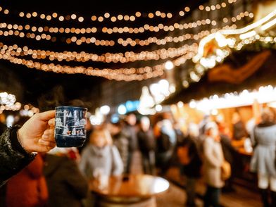 Weihnachtsdorf in Einbeck im Lichterglanz Hand, die eine Tasse Glühwein hält im Fokus und das beleuchtete Einbecker Weihnachtsdorf dahinter.