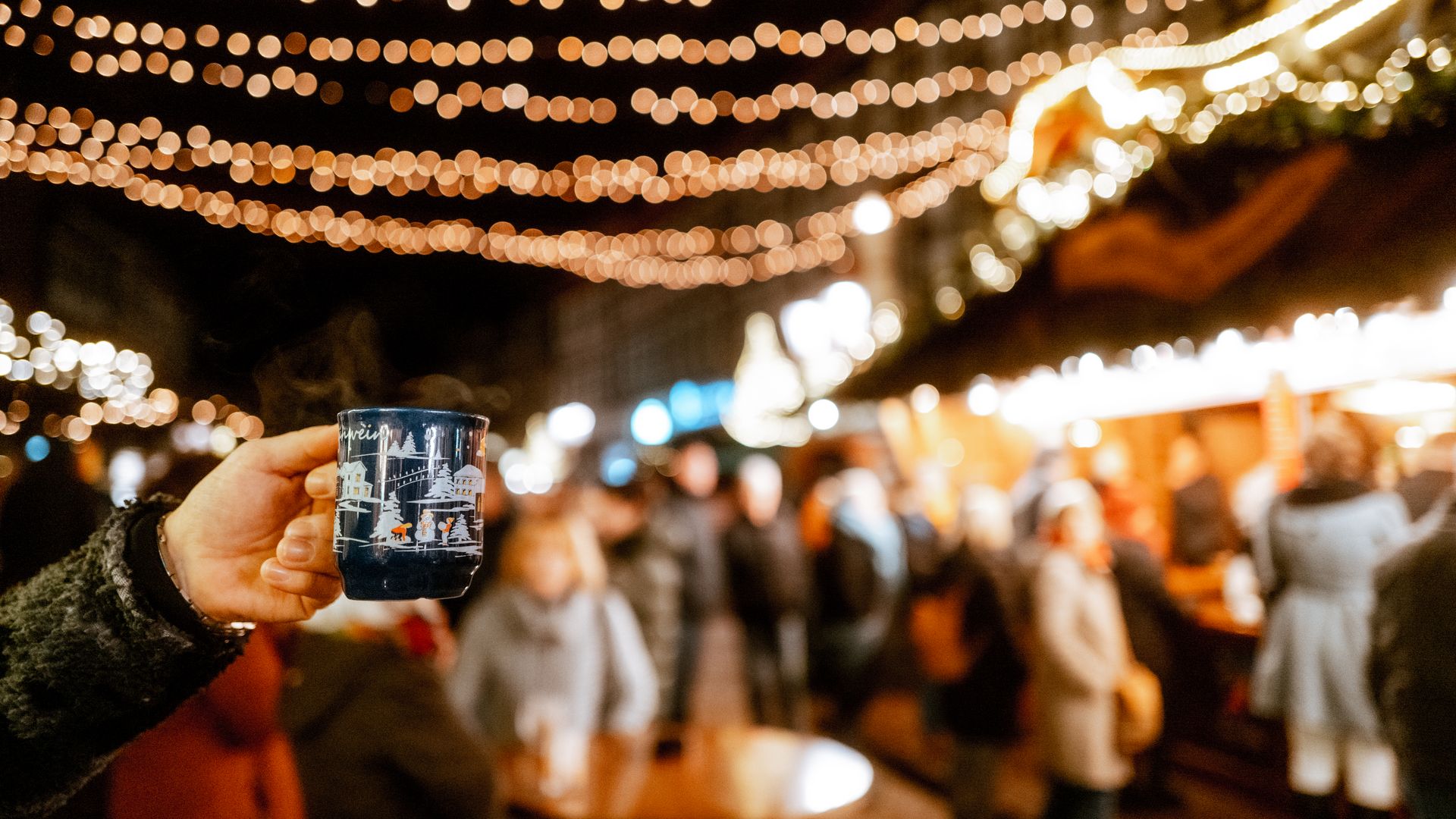 Hand, die eine Tasse Glühwein hält im Fokus und das beleuchtete Einbecker Weihnachtsdorf dahinter.