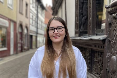 Person mit Brille steht in enger Altstadtstraße neben reich verziertem Brunnen.