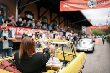 Frau fotografiert aus gelbem Oldtimer-Cabriolet die PS.SPEICHER Rallye mit Zuschauern.