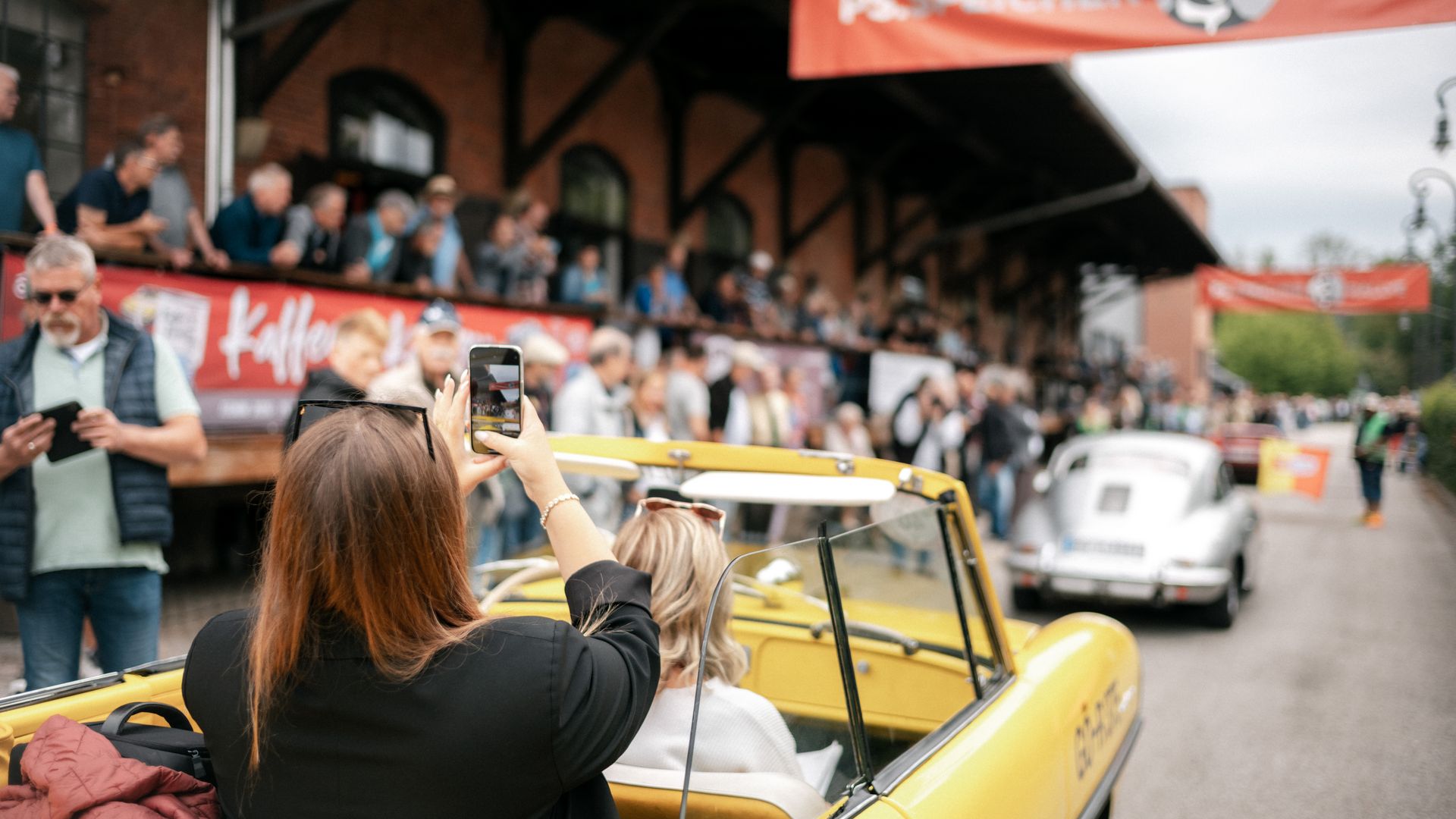 Frau fotografiert aus gelbem Oldtimer-Cabriolet die PS.SPEICHER Rallye mit Zuschauern.