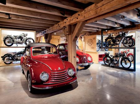 Ein roter Oldtimer steht in der hellen Ausstellung des PS.SPEICHER. Zwischen massiven Holzbalken werden im Hintergrund weitere historische Autos und alte Motorräder auf Podesten präsentiert.