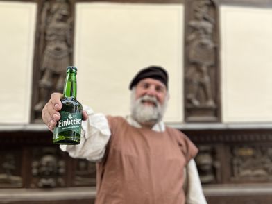 Einbecker Braumeister hält vor einem Fachwerkhaus eine Einbecker Bierflasche in die Kamera