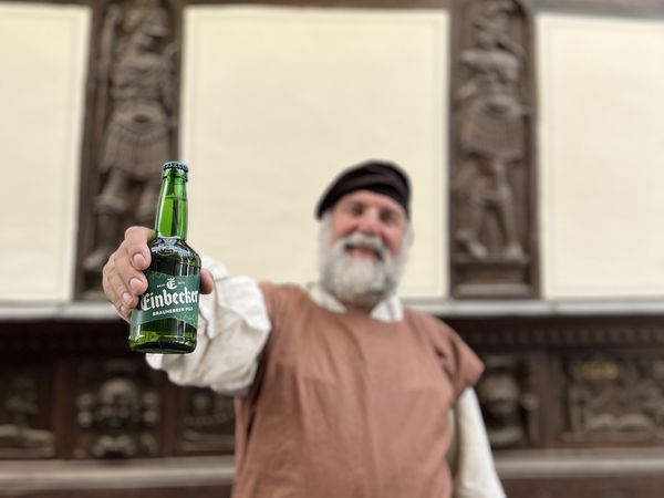 Einbecker Braumeister hält vor einem Fachwerkhaus eine Einbecker Bierflasche in die Kamera