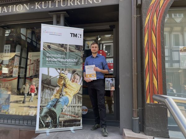 Ein Mann steht vor einer Tourist-Info, hält eine Urkunde und posiert neben einem Roll-up-Banner.