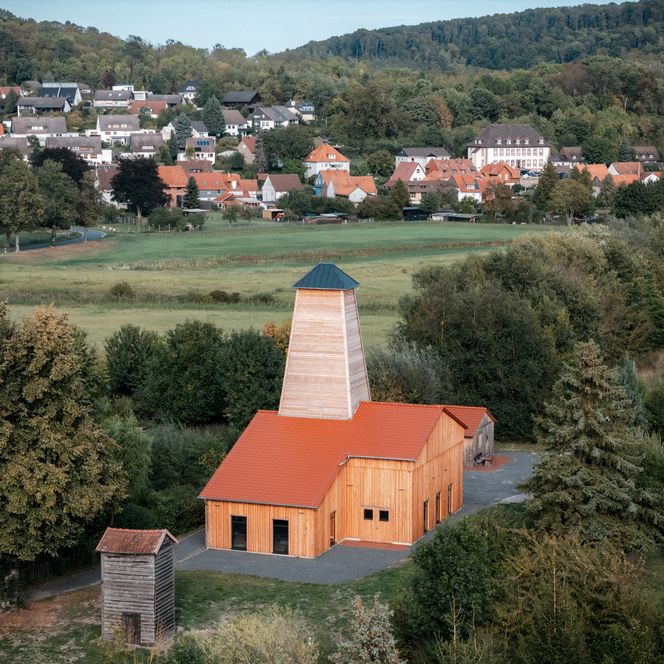 Holzverkleidete Saline mit rotem Dach vor grünen Wiesen und dem Dorf Salzderhelden im Hintergrund.
