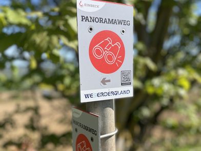 Weißes Schild für den Panoramaweg mit rotem Fernglas-Symbol und Richtungspfeil im Grünen.
