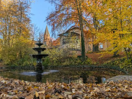 Backsteinvilla mit Brunnen im herbstlich gefärbten Park, Laub auf dem Boden und blauer Himmel.